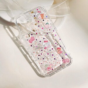 May include: Clear phone case with a pink and purple glitter design. The case has a Hello Kitty character and other small charms. The case also has the name "Jesse" written on the back.