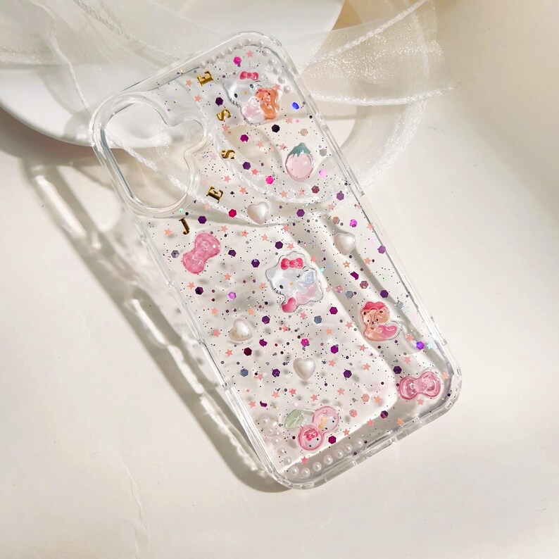 May include: Clear phone case with a pink and purple glitter design. The case has a Hello Kitty character and other small charms. The case also has the name "Jesse" written on the back.