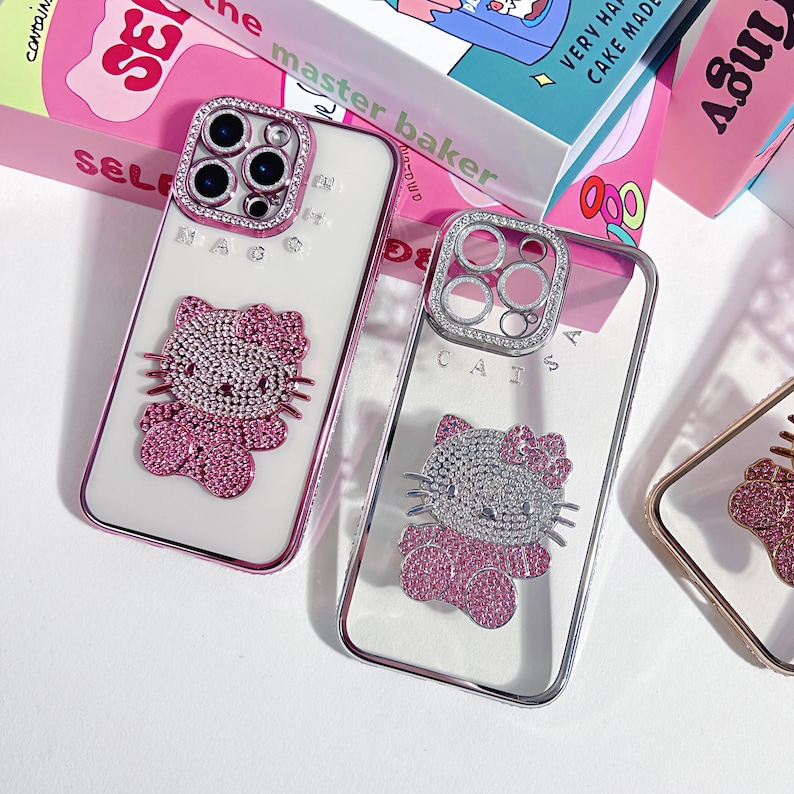 K&ouml;nnte beinhalten: Zwei transparente Handyh&uuml;llen mit einem Hello Kitty-Design. Die H&uuml;llen haben einen rosa-silbernen Rand und sind mit Strasssteinen verziert. Die H&uuml;llen haben einen Kameraschutz mit Strasssteinen. Der Text "MAG" und "CATSA" ist sichtbar.