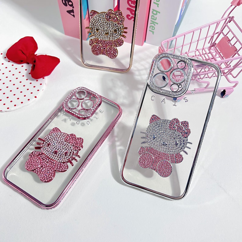 K&ouml;nnte beinhalten: Drei transparente Handyh&uuml;llen mit einem rosafarbenen Hello-Kitty-Design. Die H&uuml;llen haben ein erhabenes, mit Schmuck besetztes Hello-Kitty-Design und eine Schmuckabdeckung f&uuml;r die Kameralinse. Die H&uuml;llen befinden sich auf einer wei&szlig;en Oberfl&auml;che.