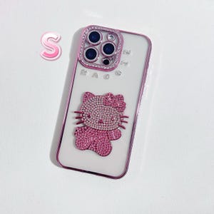 K&ouml;nnte beinhalten: Eine wei&szlig;e Handyh&uuml;lle mit einem rosafarbenen Hello-Kitty-Design und einer rosafarbenen, mit Strass besetzten Kameraobjektivabdeckung. Auf der H&uuml;lle steht "MAG".