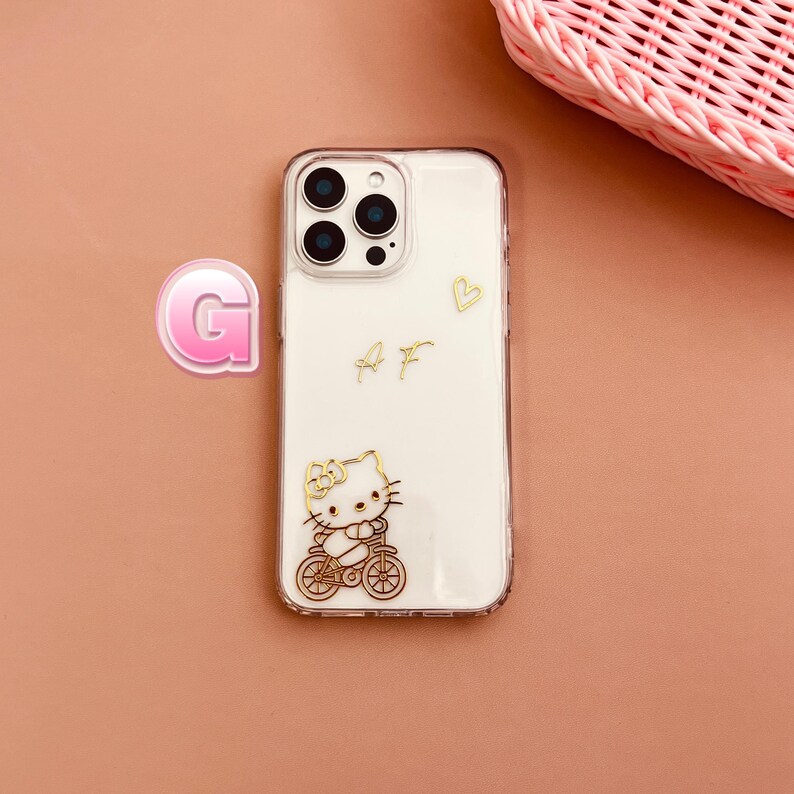May include: Clear phone case with a gold Hello Kitty design riding a bicycle. The case has a white background with gold lettering and a small gold heart. A pink letter "G" and a pink basket are in the background.
