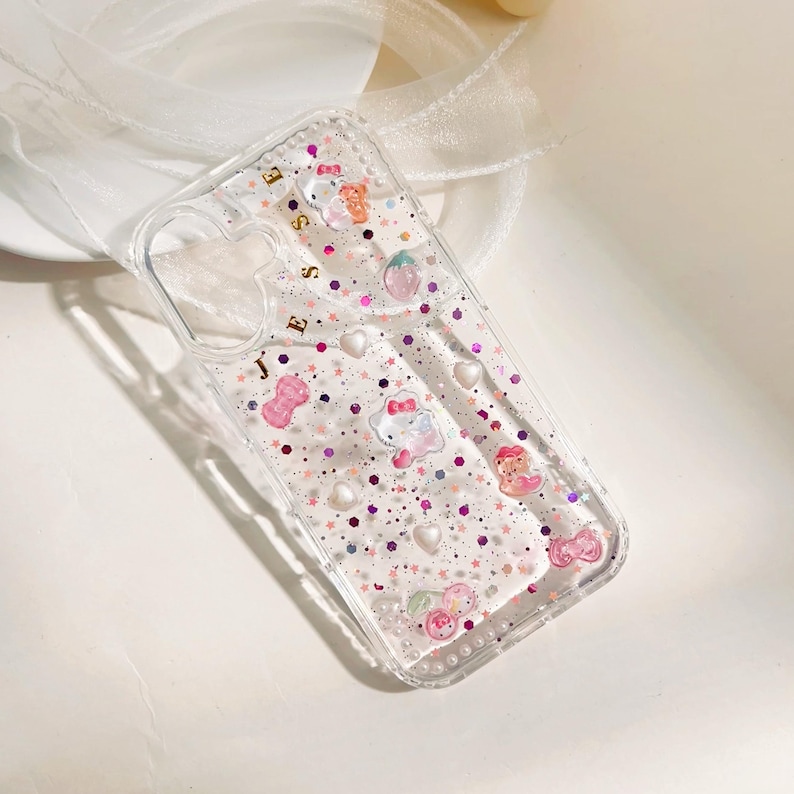 May include: Clear phone case with glitter and decorative charms. The case features pink bows, white hearts, and cartoon characters. The word "JESS" is written in gold letters on the case.