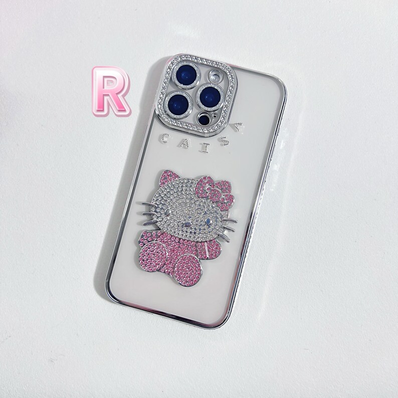 K&ouml;nnte beinhalten: Wei&szlig;es iPhone-Case mit einem Hello-Kitty-Design mit rosa und wei&szlig;en Strasssteinen. Das Case hat eine transparente Kameraobjektivabdeckung mit silbernem Rand und den Schriftzug "CAIS" auf der R&uuml;ckseite.