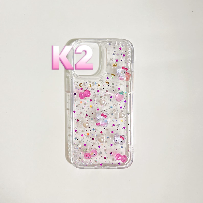 May include: Clear phone case with a glittery design. The case features pink, purple, and white glitter, along with small Hello Kitty character designs and the word "CASES". The letter "K2" is in the upper left corner.