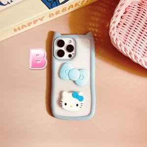 Puede incluir: Una funda de tel&eacute;fono azul claro con fondo blanco y un personaje de Hello Kitty, un lazo azul y un recorte para el objetivo de la c&aacute;mara.