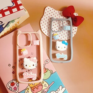 Op de afbeelding: Twee roze en grijze telefoonhoesjes met een Hello Kitty-ontwerp. De hoesjes hebben een transparante achterkant en een roze en grijze rand. De hoesjes hebben een Hello Kitty-ontwerp met de woorden "Baby" onderaan. De hoesjes liggen op een roze en blauwe achtergrond met een wit stippenpatroon.