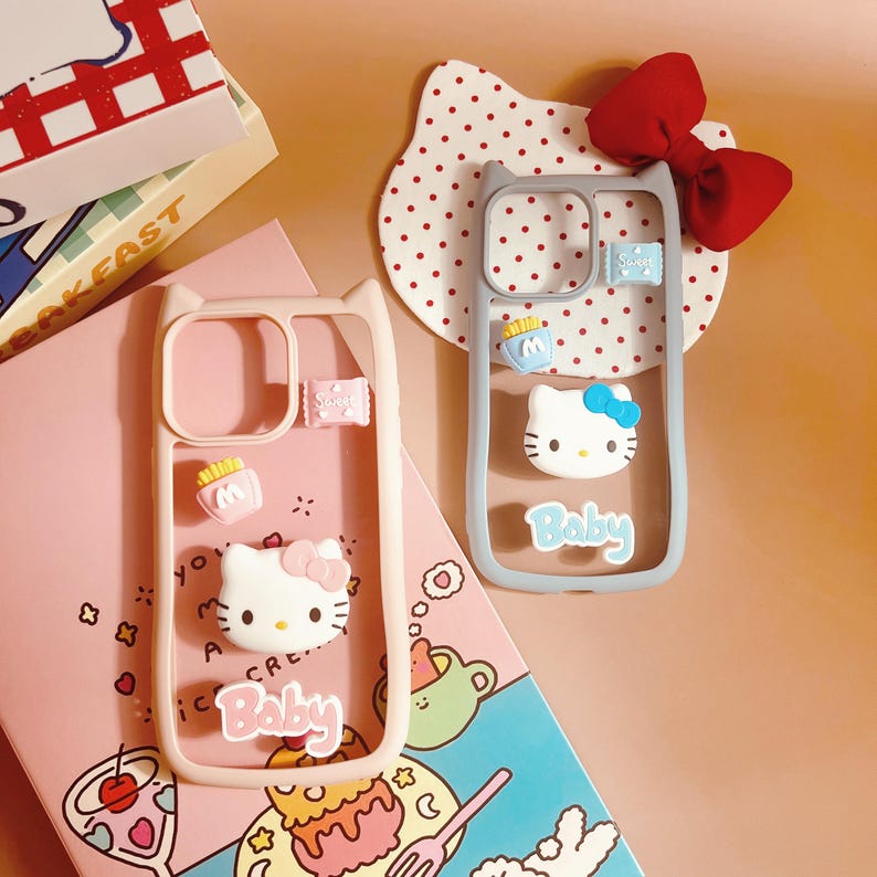 Op de afbeelding: Twee roze en grijze telefoonhoesjes met een Hello Kitty-ontwerp. De hoesjes hebben een transparante achterkant en een roze en grijze rand. De hoesjes hebben een Hello Kitty-ontwerp met de woorden "Baby" onderaan. De hoesjes liggen op een roze en blauwe achtergrond met een wit stippenpatroon.