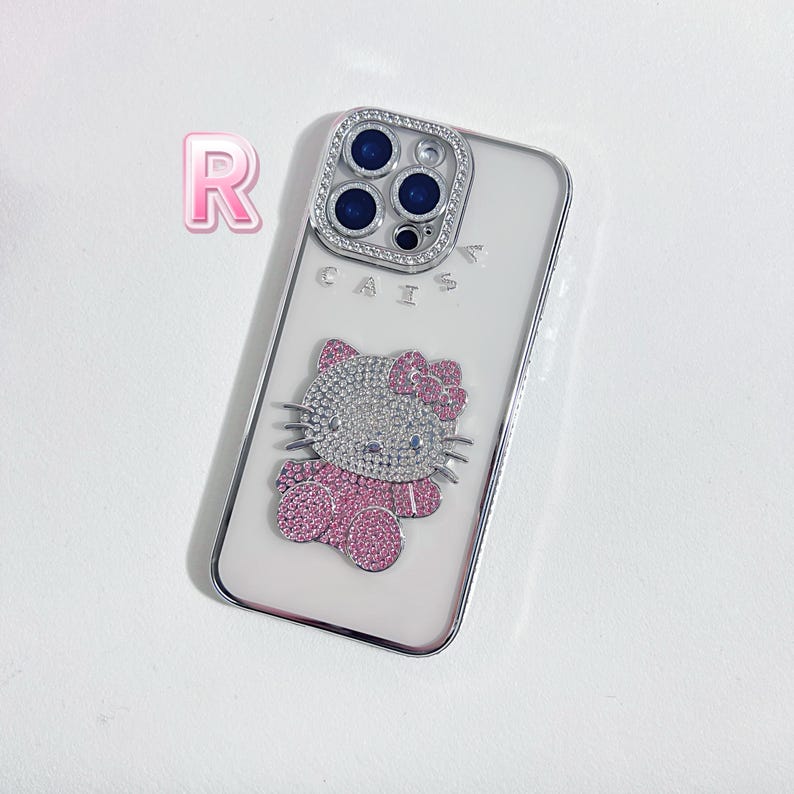 Pu&ograve; includere: Custodia bianca per iPhone con un design Hello Kitty con strass rosa e bianchi. La custodia ha una copertura trasparente per l'obiettivo della fotocamera con un bordo argentato e la parola "CAIS" stampata sul retro.