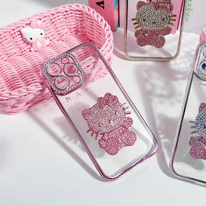 K&ouml;nnte beinhalten: Eine transparente Handyh&uuml;lle mit einem rosafarbenen Hello-Kitty-Design und Strasssteinen. Die H&uuml;lle hat einen Ringhalter mit einem transparenten Hintergrund und Strasssteinen. Die H&uuml;lle liegt auf einem rosafarbenen Weidenkorb.