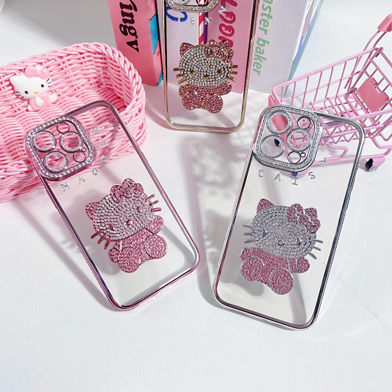 May include: Three clear phone cases with pink and silver Hello Kitty designs. Each case has a different design of Hello Kitty with rhinestones. The cases are on a pink surface. The text on the cases reads 'CAIS' and 'MAO'.