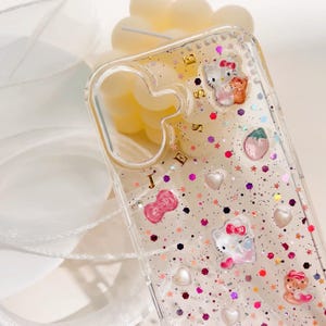 May include: A clear phone case with a glittery background and Hello Kitty characters. The case has a gold inscription that reads "JESSIE".