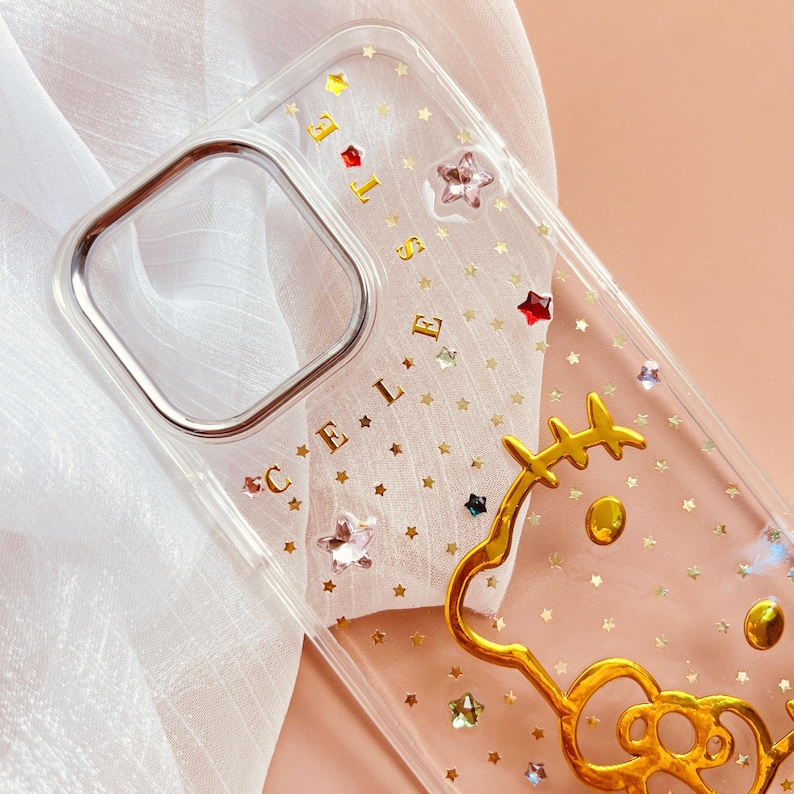 May include: Clear phone case with a gold glitter background and a gold Hello Kitty outline. The case has gold stars and rhinestones.