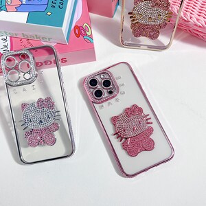 K&ouml;nnte beinhalten: Transparente Handyh&uuml;llen mit einem Hello Kitty-Design. Die H&uuml;llen zeigen ein rosa-silbernes Strass-Design der ikonischen Figur. Die H&uuml;llen haben eine transparente R&uuml;ckseite und einen silbernen oder rosa Rahmen. Auf einer wei&szlig;en Oberfl&auml;che.