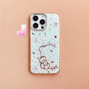 Pu&ograve; includere: Custodia per telefono bianca con un design Hello Kitty argentato e uno sfondo scintillante. La custodia ha un retro trasparente e un bordo argentato.