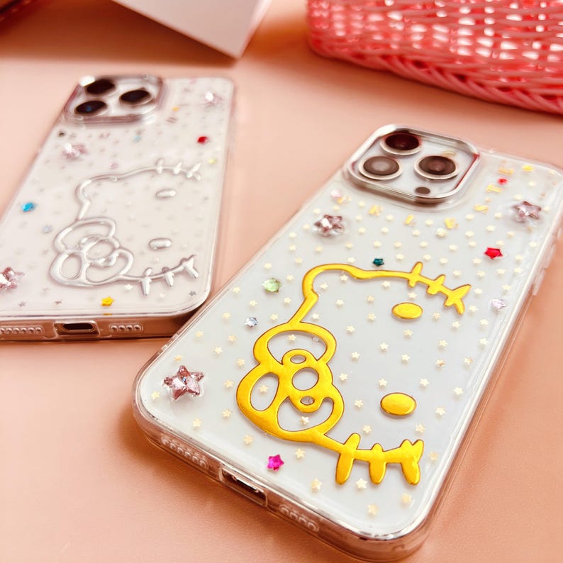 以下が含まれることがあります： 白い背景と金色のグリッターが付いた透明な携帯電話ケースが2つ。1つのケースは銀色のハローキティの輪郭、もう1つのケースは金色のハローキティの輪郭です。
