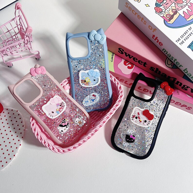 Może przedstawiać: Trzy błyszczące etui na telefon z motywami Hello Kitty. Jedno jest r&oacute;żowe, jedno niebieskie, a jedno czarne. Każde etui ma kokardkę i detale uszu kota. Etui znajdują się w r&oacute;żowym koszyku, z książkami w tle.