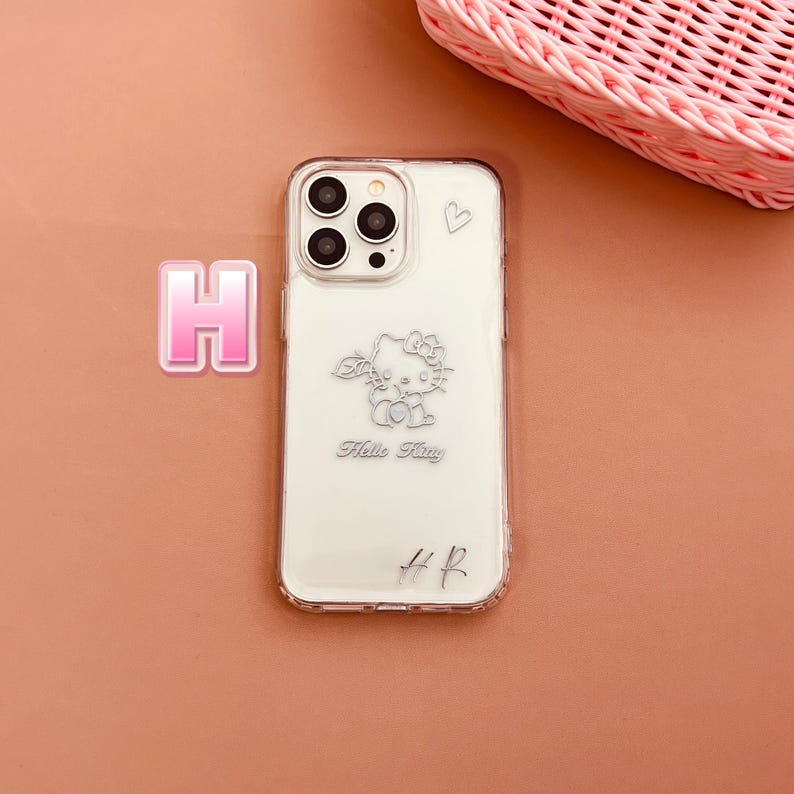 May include: Clear phone case with a Hello Kitty design. The case features a silver outline of Hello Kitty, a small heart, and the text "Hello Kitty". The case also has the initials "H R" at the bottom.