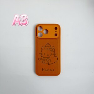 Puede incluir: Funda de tel&eacute;fono naranja con un dise&ntilde;o de gato de dibujos animados y el nombre "Hanna" grabado en la parte inferior. La funda tiene un recorte de c&aacute;mara naranja y un cuerpo marr&oacute;n claro. La imagen tambi&eacute;n muestra el texto "A3" en rosa.