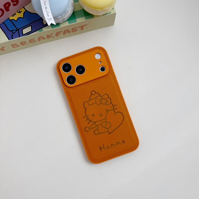 May include: Orange phone case featuring a Hello Kitty design and the name "Hanna". The case has a camera cutout and a textured finish. The case is designed for a smartphone.