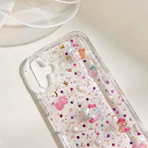May include: A clear phone case with a glittery pink and purple background. The case has a gold inscription that says "Jesse" and features various cartoon characters, including Hello Kitty, a strawberry, and a bow.