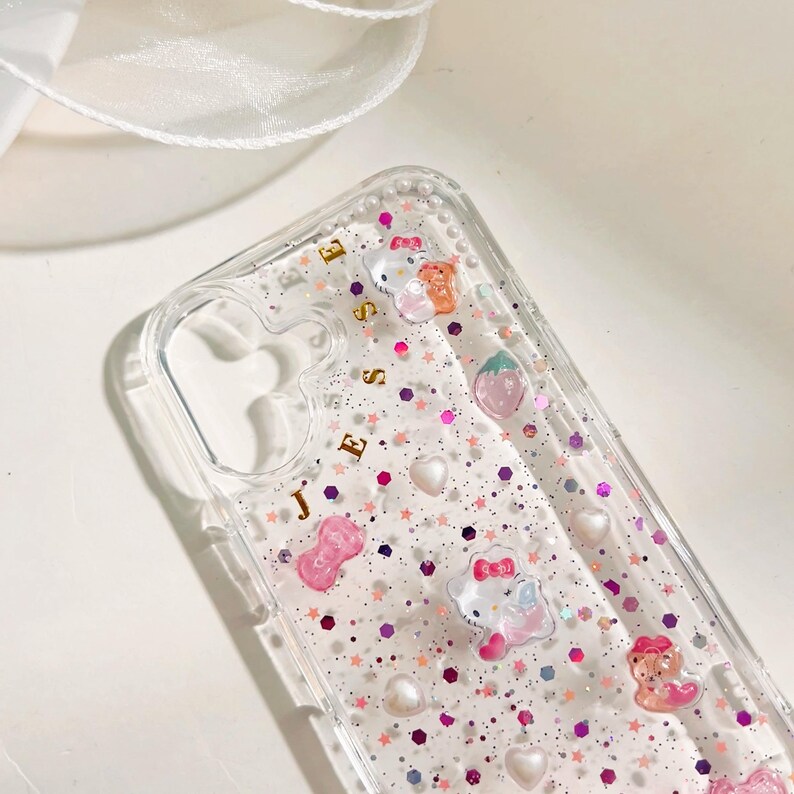 May include: A clear phone case with a glittery pink and purple background. The case has a gold inscription that says "Jesse" and features various cartoon characters, including Hello Kitty, a strawberry, and a bow.