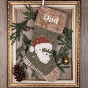 May include: A Christmas stocking featuring a textured olive green body with a plush tan cuff embroidered with the word Dad in white. The stocking displays a central Santa Claus applique wearing a red hat and includes a plaid heel and toe.