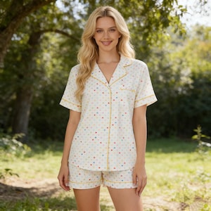 May include: A two-piece pajama set featuring a short-sleeved button-down shirt and matching shorts. The set is cream-colored with a pattern of small, colorful hearts. The edges are trimmed with yellow piping.