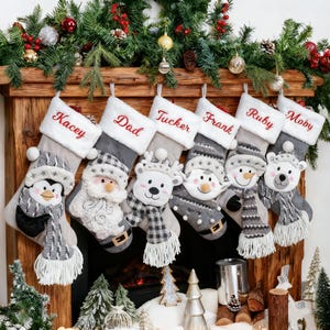 May include: A collection of Christmas stockings hanging above a fireplace. Each stocking is gray and white, with a plush animal face and a name embroidered in red. The animals include a penguin, Santa, a reindeer, a snowman, and a polar bear.