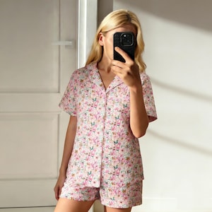 May include: A two-piece pajama set featuring a pink floral pattern. The set includes a short-sleeved button-down shirt and matching shorts. The floral design incorporates shades of pink, white, and blue.