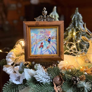 May include: A small framed painting depicting a winter scene with two figures skiing down a snowy hill. The painting is in a brown wooden frame with a decorative metal top. The artwork is displayed on a decorative arrangement of pine, white flowers, and fairy lights.