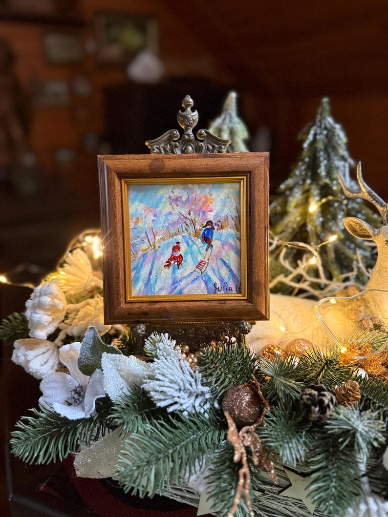 May include: A small framed painting depicting a winter scene with two figures skiing down a snowy hill. The painting is in a brown wooden frame with a decorative metal top. The artwork is displayed on a decorative arrangement of pine, white flowers, and fairy lights.