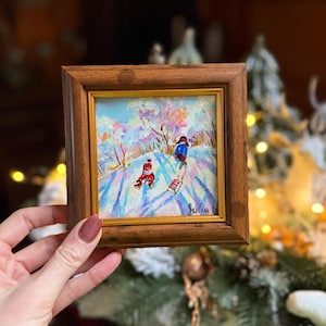 May include: A small, framed oil painting depicting a winter scene with two figures on skis. The artwork features a snowy landscape with trees and a blue sky. The frame is made of dark wood. Signed "Julia N."
