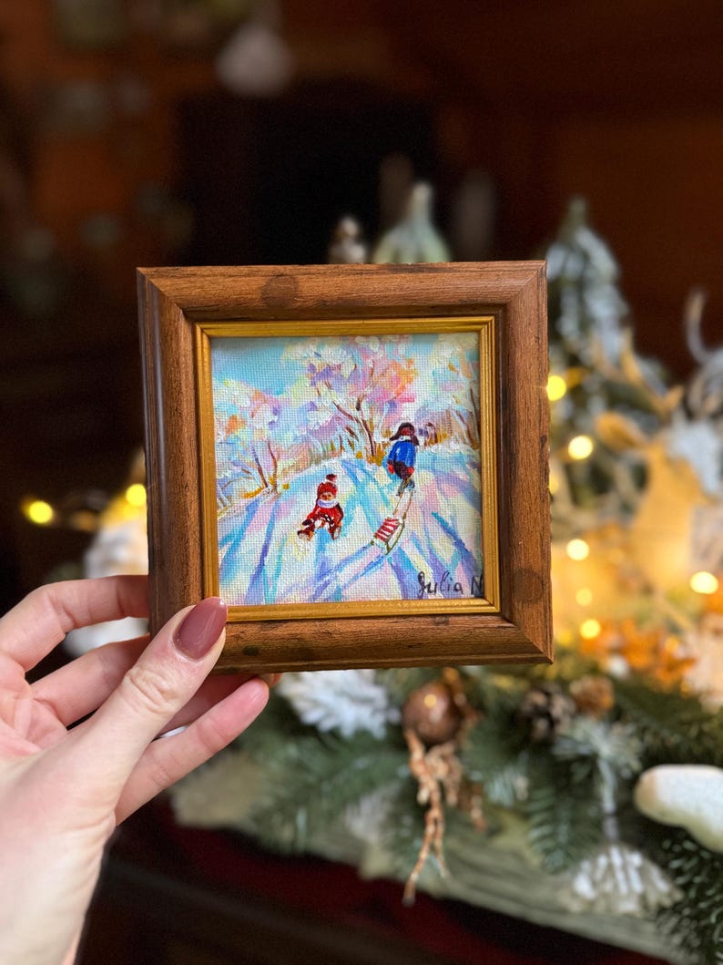 May include: A small, framed oil painting depicting a winter scene with two figures on skis. The artwork features a snowy landscape with trees and a blue sky. The frame is made of dark wood. Signed "Julia N."