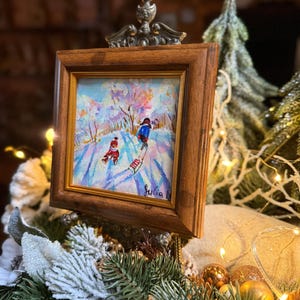 May include: A framed painting depicts a winter scene with two figures sledding down a snow-covered hill. The artwork is set in a wooden frame with a decorative metal top. The painting features a colorful landscape with a snowy environment.