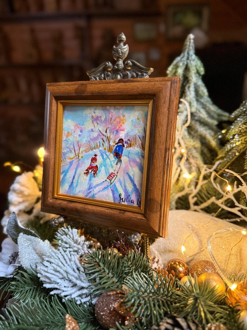 May include: A framed painting depicts a winter scene with two figures sledding down a snow-covered hill. The artwork is set in a wooden frame with a decorative metal top. The painting features a colorful landscape with a snowy environment.
