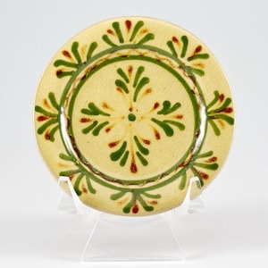 May include: A small, round ceramic plate with a cream-colored base. The plate features a green and brown circular design with green and red floral patterns. The plate is displayed on a clear plastic stand.