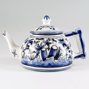 Puede incluir: Una tetera de cerámica blanca con diseños florales y paisajísticos azules. La tetera tiene un pico curvo, un asa y una tapa con un pequeño pomo. El diseño incluye hojas estilizadas y una escena acuática.