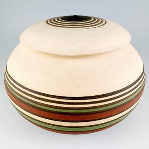 May include: A white ceramic jar with a lid, decorated with brown, red, and green stripes. The lid has a concentric circle design in brown and black.