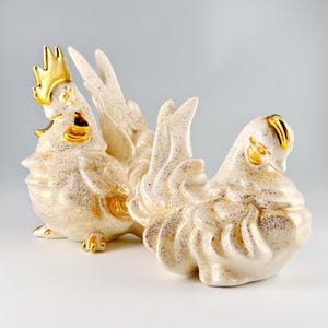 May include: Two ceramic rooster and hen figurines. The rooster stands upright with a golden comb and accents, while the hen is in a seated position. Both are off-white with speckled detailing and gold embellishments.