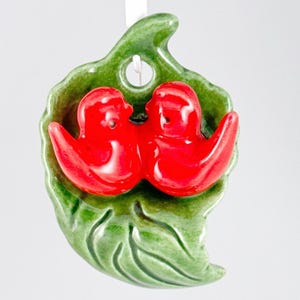 May include: A ceramic ornament featuring two red birds nestled on a green leaf. The birds have a glossy finish, and the leaf has detailed veining. A small hole is at the top for hanging.