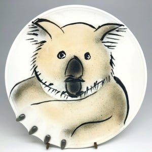 Può includere: Un piatto in ceramica bianca con un'illustrazione in bianco e nero di un koala. Il koala è disegnato in modo semplice e stilizzato, con la sua pelliccia rappresentata da una serie di brevi linee nere. Il piatto ha un bordo leggermente irregolare e tre piccoli piedini in ceramica grigi sul fondo.