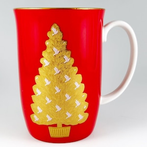 May include: A red ceramic mug with a white handle features a gold Christmas tree design with white birds. The tree is in a gold pot.