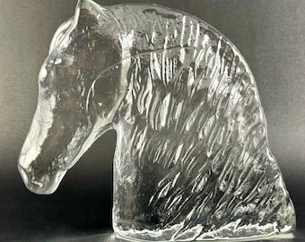 May include: Clear glass sculpture of a horse's head. The sculpture is detailed with textured lines to represent the mane and facial features. The horse head is a decorative item.