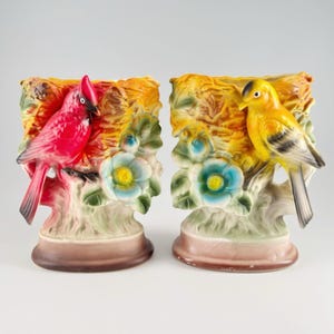 May include: Two ceramic bird-themed planters featuring a red cardinal on the left and a yellow bird on the right. Each planter is shaped like a textured nest with light blue flowers and green leaves, set upon a smooth, rounded brown base.
