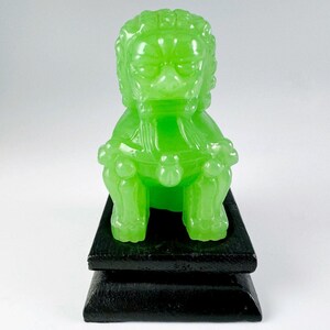 May include: A green jade foo dog figurine sitting on a black wooden base. The foo dog is a traditional Chinese guardian lion.