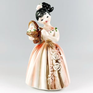 May include: A vintage ceramic figurine of a young woman holding a basket of flowers. The figurine has black hair, rosy cheeks, and is wearing a long, flowing dress with rose accents. The dress is primarily cream and brown with a red gradient.