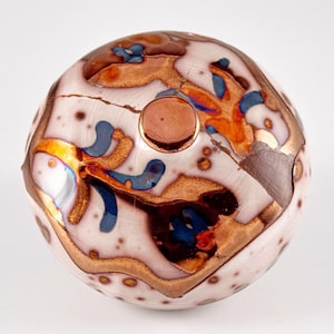 May include: A decorative, spherical object with a white base, adorned with copper-colored, blue, and orange abstract designs. A copper-colored circular element is centered on the top. The surface has a speckled appearance.