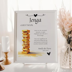Puede incluir: Un cartel enmarcado blanco con las palabras "Jenga Guest Book" impresas en negro. El cartel presenta una ilustración de acuarela de una torre de Jenga y el texto "Laissez un vœu, une pensée amusante, ou signez simplement votre nom sur ce bloc de Jenga. Nous nous rappellerons de cette journée chaque fois que nous jouerons à ce jeu lors de nos futurs moments ensemble. Amelie & Pierre 30.07.2027"
