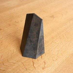 May include: A dark gray, geometric sculpture with a textured surface. The angular form is a truncated pyramid shape, resting on a light brown wooden surface. The sculpture's height is approximately 6 inches.